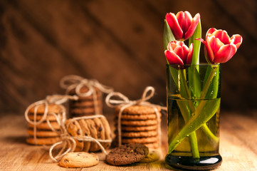 Fototapeta na wymiar Simple rustic still life of red tulips in a green glass and stacks various cookies on wooden table.