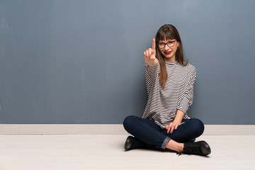 Woman with glasses sitting on the floor showing and lifting a finger