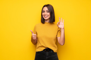 Young woman over yellow wall showing ok sign with and giving a thumb up gesture - obrazy, fototapety, plakaty