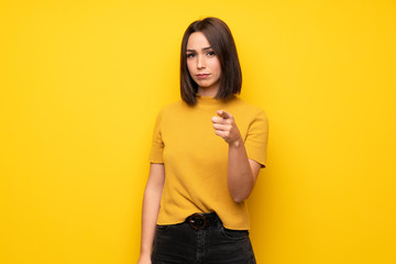 Young woman over yellow wall frustrated and pointing to the front