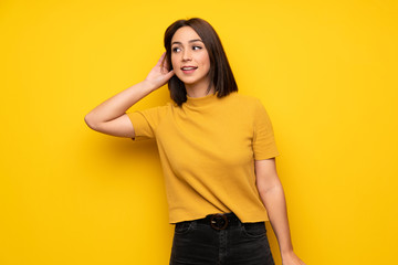 Young woman over yellow wall listening to something by putting hand on the ear