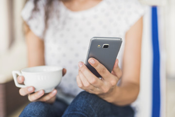 Young happy woman using smartphone at home. Girl holding mobile phone and coffee cup