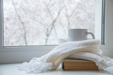 Mug of hot tea and warm woolen knitting on windowsill against snow landscape from outside.
