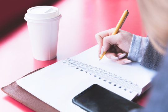 Close-up From Behind The Back Of A Young Woman In A Winter Polto Is Holding A Ballpoint Pen And Is Going To Write Something Down In A Notebook On A Red Table. Notes.