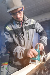 Young carpenter with a beard working with an electric plane without suction of sawdust. Leveling and sanding wooden bars. Low wide angle