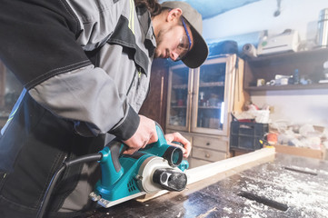 Young carpenter with a beard working with an electric plane without suction of sawdust. Leveling and sanding wooden bars. Low wide angle