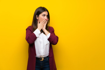 Young woman over yellow wall covering mouth and looking to the side