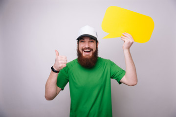 Smiling bearded man with speech bubble makes ok sign while standing on light gray background, portrait picture