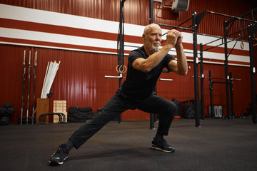 Attractive self determined elderly male with gray hair and beard holding hands in front of him while doing side lunges in gym, having serious concentrated facial expression, dressed in sportswear