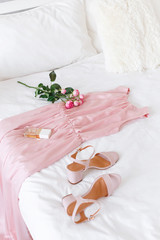 Obraz na płótnie Canvas Elegant long pink dress, heeled sandals, perfume and rose flowers lying on the bed with white linen. Girl choosing outfit. White female modern stylish bedroom.