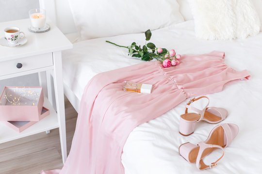 Elegant long pink dress, heeled sandals, perfume and rose flowers lying on the bed with white linen. Girl choosing outfit. White female modern stylish bedroom.