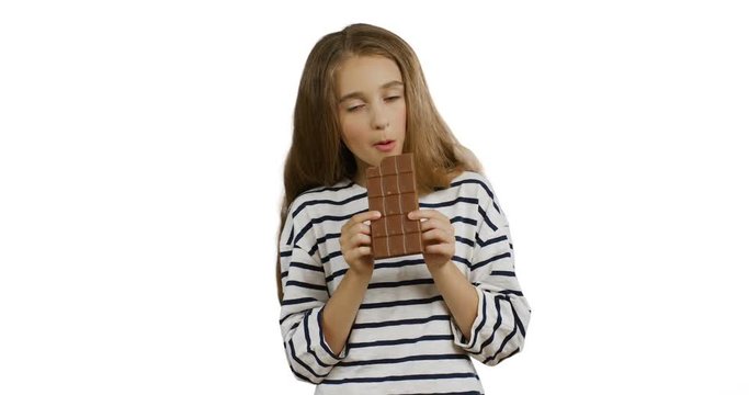 Cute blonde teen girl in the striped blouse holding a bar of chocolate, biting it and eating it on the white background.
