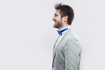 Profile side view portrait of happy satisfied handsome bearded man in casual grey suit and blue bow tie standing and looking straight with toothy smile. studio shot, isolated on light grey background.