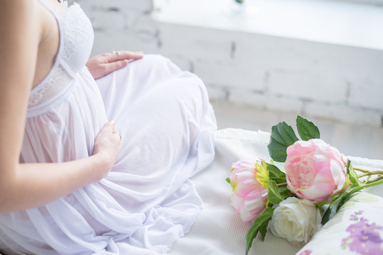 Close-up Image of pregnant woman in nice white dress touching her belly with hands and holding a bouquet of peonies. Beautiful image of a pregnant girl with flowers. Expecting a child. Motherhood.