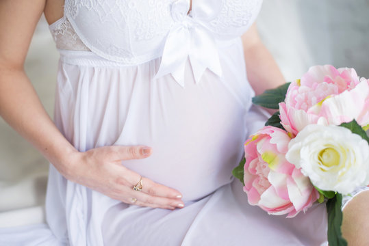 Close-up Image of pregnant woman in nice white dress touching her belly with hands and holding a bouquet of peonies. Beautiful image of a pregnant girl with flowers. Expecting a child. Motherhood.