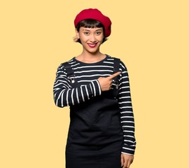 Young woman with beret pointing to the side to present a product over yellow background
