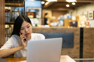 Happy young Asian woman using phone and working with a laptop in the coffee shop cafe office.