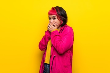 Young woman with pink hair over yellow wall covering mouth and looking to the side