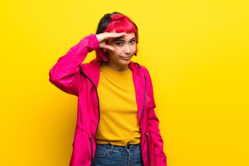 Young woman with pink hair over yellow wall looking far away with hand to look something
