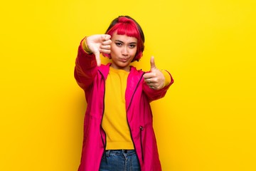 Young woman with pink hair over yellow wall making good-bad sign. Undecided between yes or not