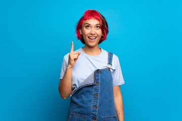 Young woman with pink hair over blue wall pointing up and surprised