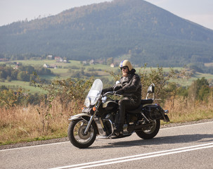 Bearded motorcyclist in helmet, sunglasses and black leather clothing riding powerful cruiser motorcycle down sunny asphalt road on bright summer day on background of green woody hill