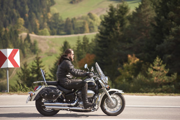 Obraz na płótnie Canvas Side view of bearded long-haired motorcyclist in sunglasses and black leather clothing riding cruiser motorbike along narrow asphalt path on sunny day on background of tall trees and greeen grass.