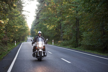 Bearded biker in sunglasses, helmet and black leather clothing riding cruiser motorcycle along asphalt road winding among tall green trees on sunny summer evening.