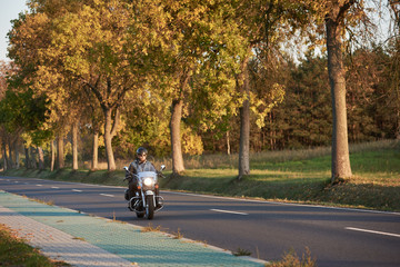 Bearded biker in sunglasses and black leather clothing riding modern powerful motorcycle along empty asphalt country road by beautiful tall trees with golden foliage on sunny autumn day.