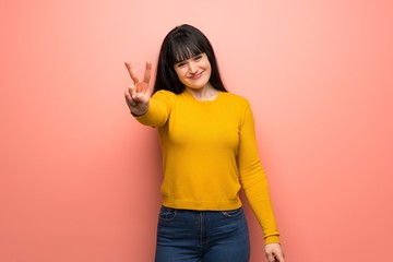 Woman with yellow sweater over pink wall smiling and showing victory sign