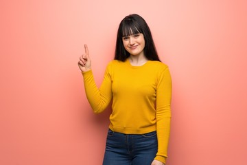 Woman with yellow sweater over pink wall showing and lifting a finger in sign of the best