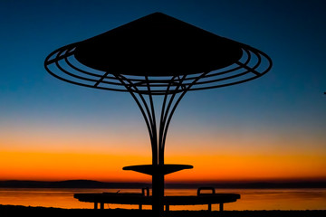 Fototapeta na wymiar Incredible, bright sunset over the water against the background of which the silhouette of a beach umbrella with a bench, a gazebo. Minsk Sea, Belarus