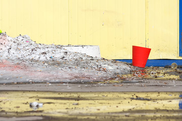 Red plastic bucket against a yellow wall with blue details, next to cigarette butts and a snowdrift of dirty snow.
