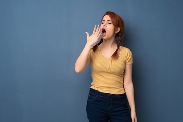 Young redhead woman over blue background yawning and covering wide open mouth with hand