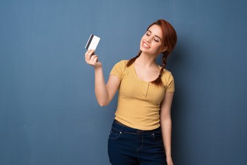 Naklejka premium Young redhead woman over blue background holding a credit card and thinking