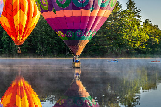 An image of hot air balloons dancing on the water of a calm misty river in the early morning sunshine.