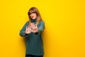 Woman with glasses over yellow wall is a little bit nervous and scared stretching hands to the front