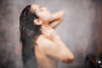 Young attractive sexy woman in shower. Blurred photo. Water wapor on glass wall. Dark-haired model stand under water stream and wash body. Enjoyment.