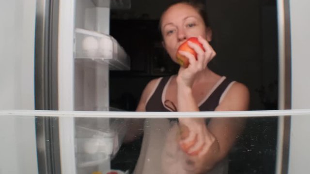 girl opens refrigerator at night, takes apple and bites it. Closeup.