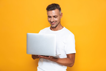 Image of attractive guy 30s in white t-shirt holding and using silver laptop