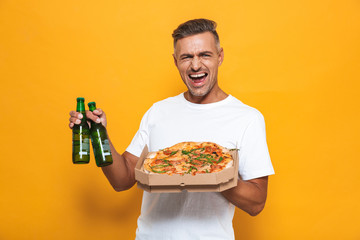 Image of cheerful man 30s in white t-shirt drinking beer and eating pizza while standing isolated