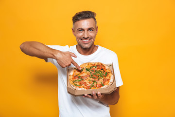 Image of optimistic man 30s in white t-shirt holding and eating pizza while standing isolated
