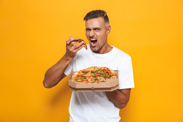 Image of caucasian man 30s in white t-shirt holding and eating pizza while standing isolated