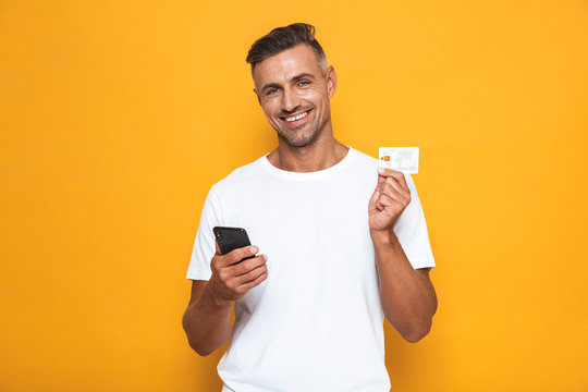 Image of cheerful guy 30s in white t-shirt holding mobile phone and credit card