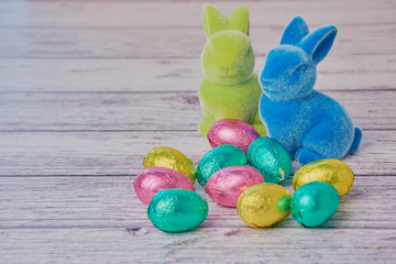 two colored easter bunnies and colored wrapped chocolate eggs beside on a white wooden background with copy space