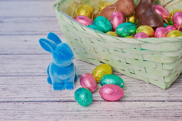 blue easter bunny beside colored chocolate eggs, in the background an easter basket on white wooden background