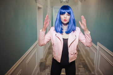 Fashion freak. Glamour synthetic girl, fake doll with blue hair is looking at the camera while standing in long corridor. Stylish woman in pink jacket in the house. Fashion and beauty concept