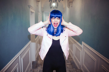 Fashion freak. Glamour synthetic girl, fake doll with blue hair is screaming and holding her head in long corridor. Stylish woman in pink jacket in the house. Fashion and beauty concept