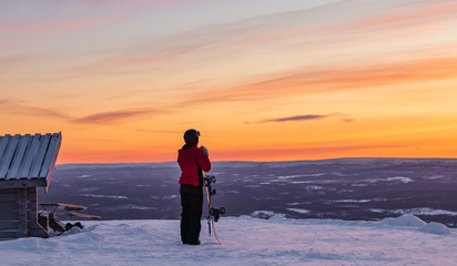 Woman standing on top of Idre Fjall, Sweden with her snowboard above her head watching a dramatic sunset over the valley below with frozen forest and mountains in the distance. 