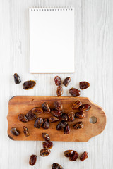 Top view, dried dates on rustic wooden board, blank notepad on a white wooden surface. From above, overhead, flat lay. Space for text.